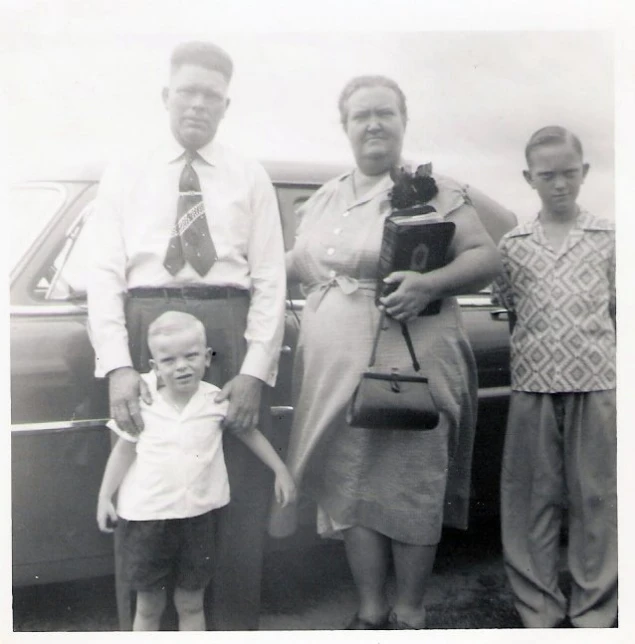 two adults and two children stand beside an old car