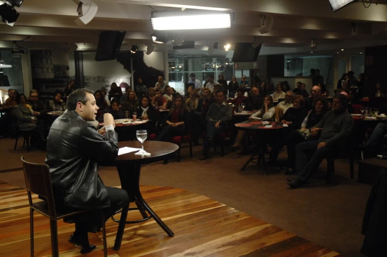 man in leather jacket sitting at table with microphone in room full of people