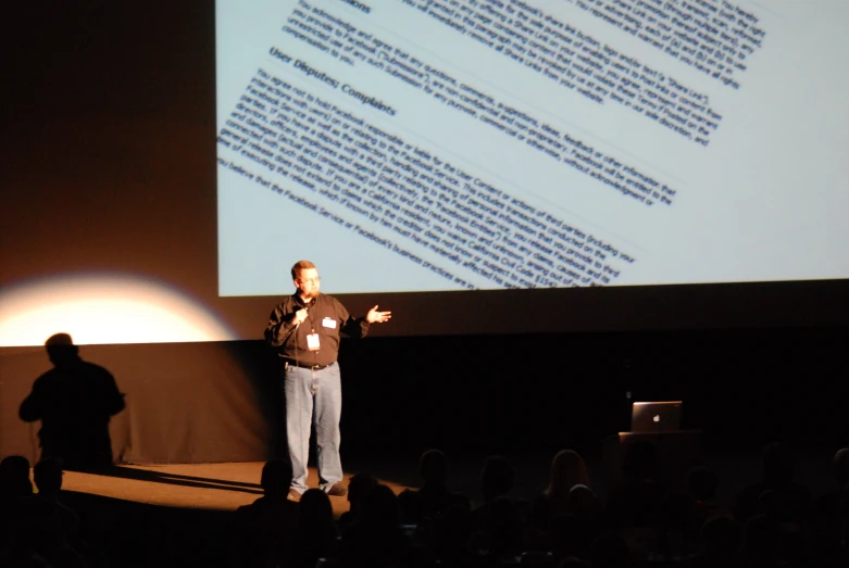 man standing in front of a projection screen showing what is written on the back wall