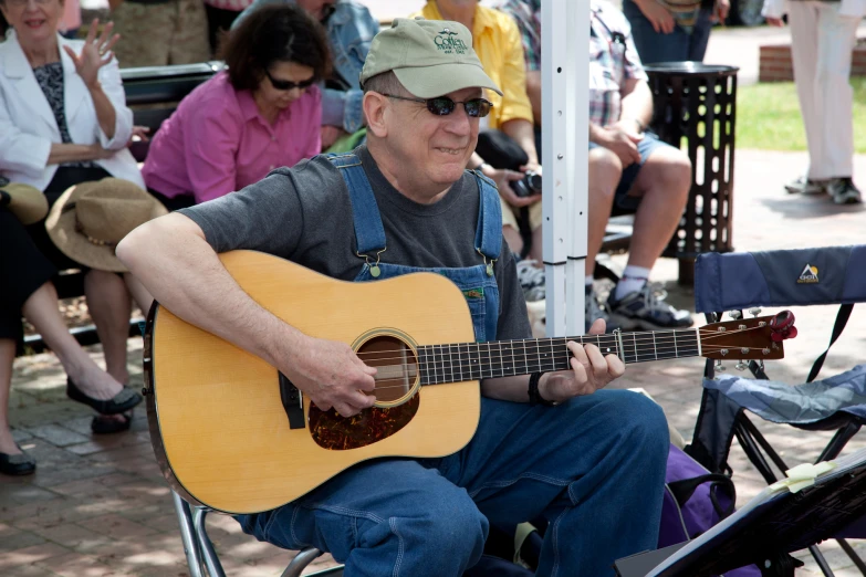 a man with a hat and suspenders sitting and playing an acoustic guitar