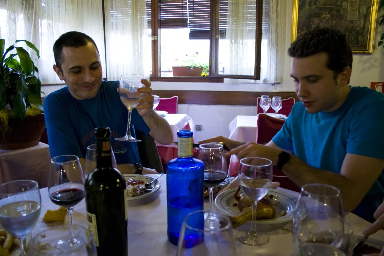 two men sitting at a table drinking wine, and one man looking down while eating