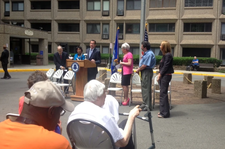 several people on the sidewalk with microphones in front of some buildings