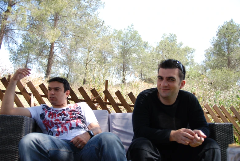 two guys sitting on a sofa outside with trees in the background