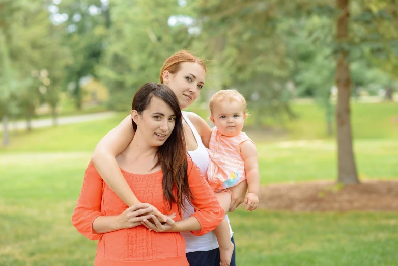 a woman holding a baby and wearing an orange top stands in the park with another woman