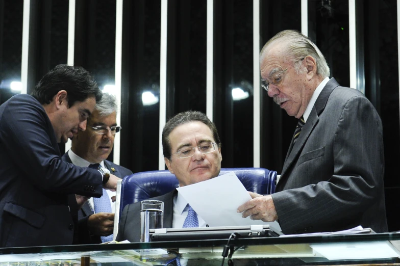 three men in suits standing at a table looking at papers