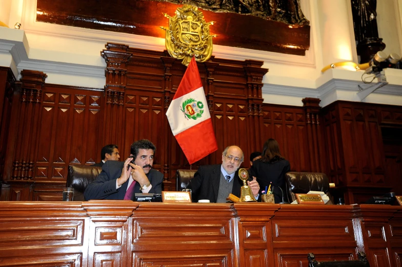 two men sitting at a wooden table in front of a flag