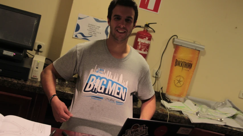 man in grey shirt standing inside his living room with laptop