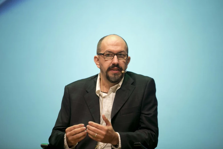 man with glasses sitting down and talking at the same time