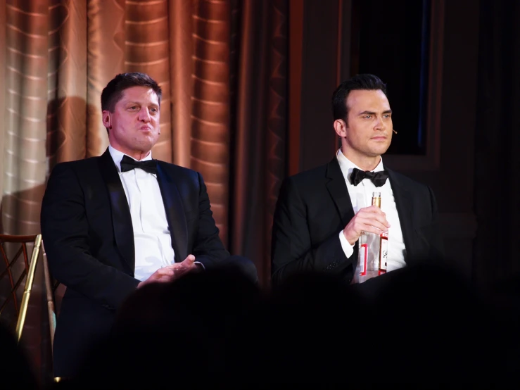 a man in black suit sitting on a chair with another man in white shirt and bow tie and other person