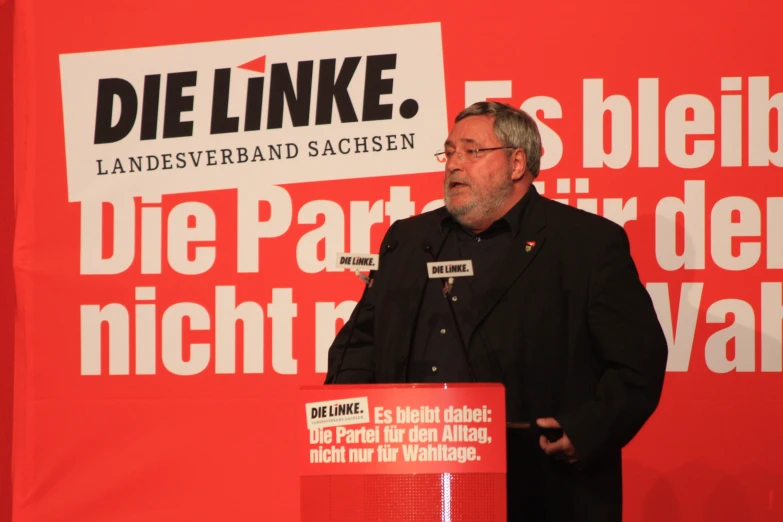 man wearing black standing at a podium in front of a red background