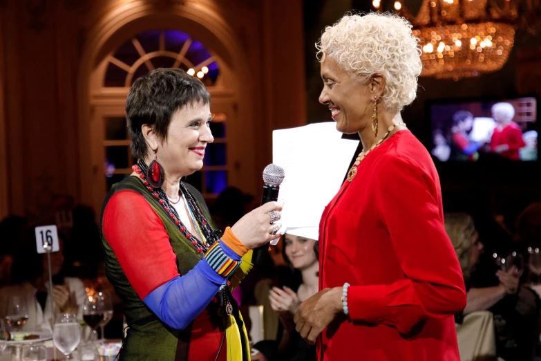 a woman in a red suit holds a microphone to a white sheet while another woman stands near her