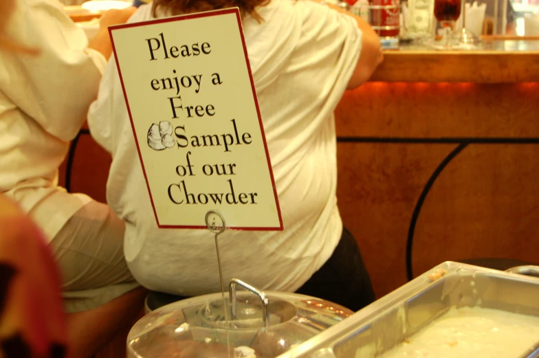 the pregnant woman is sitting in front of the stand with a sign saying, please enjoy a free sample of our chowder