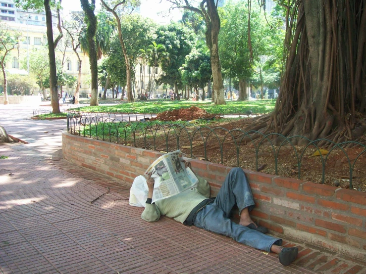 a man in jeans laying on the sidewalk reading a newspaper