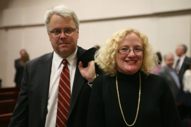 a woman with glasses in a black shirt and gold chain and a man in suit with a black jacket are walking together