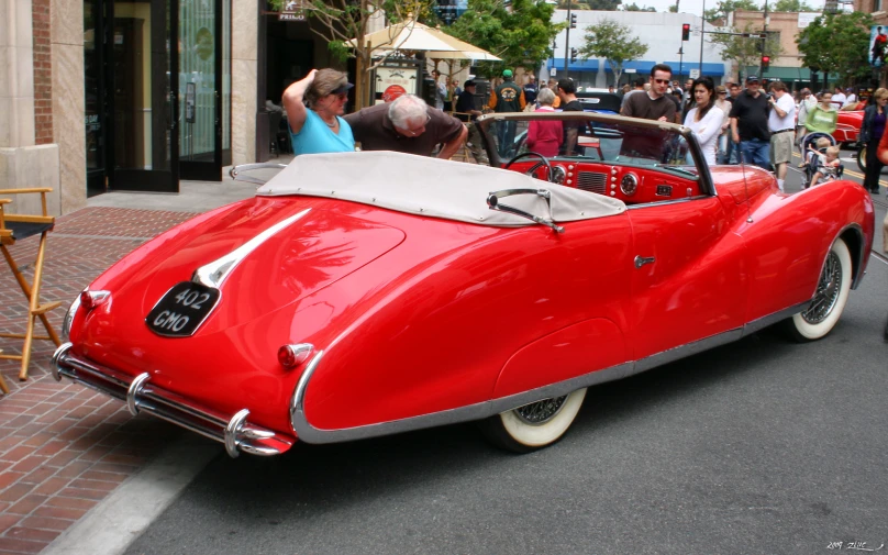 a classic car parked in the street with people and shops on the side