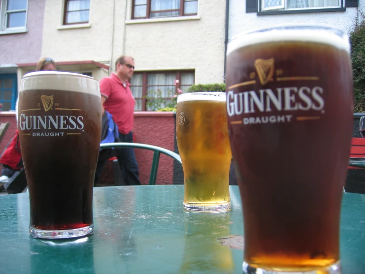 three glasses of beer sit on a table outside a pub