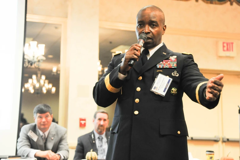 man in uniform speaking into a microphone with other people in the background