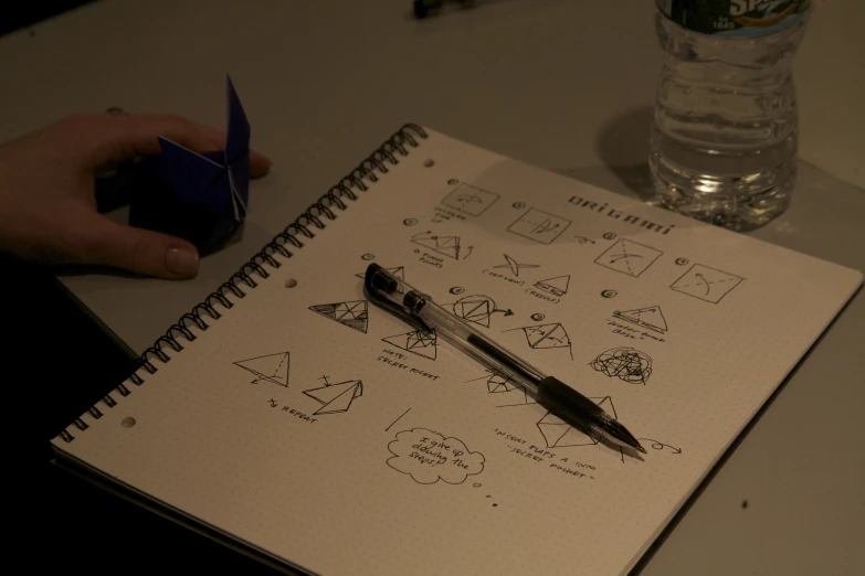 a person's hand holding a pen next to a notebook that contains sketches of things