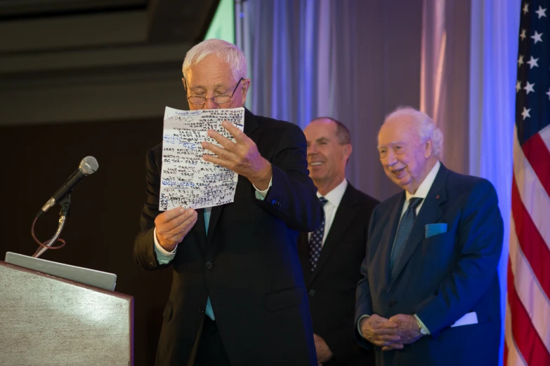 a man stands at a podium while two other men look on