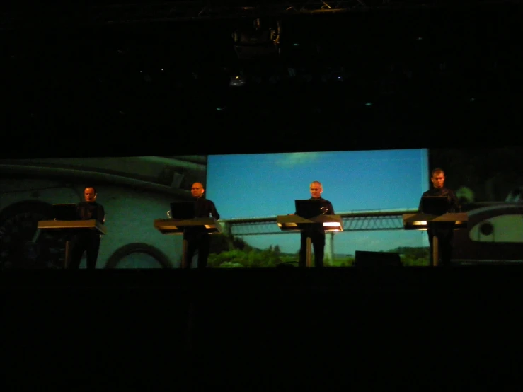 three people standing on stage while one person is playing his instrument and another has his laptop on