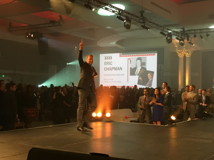 man standing on stage giving a speech with crowd behind him