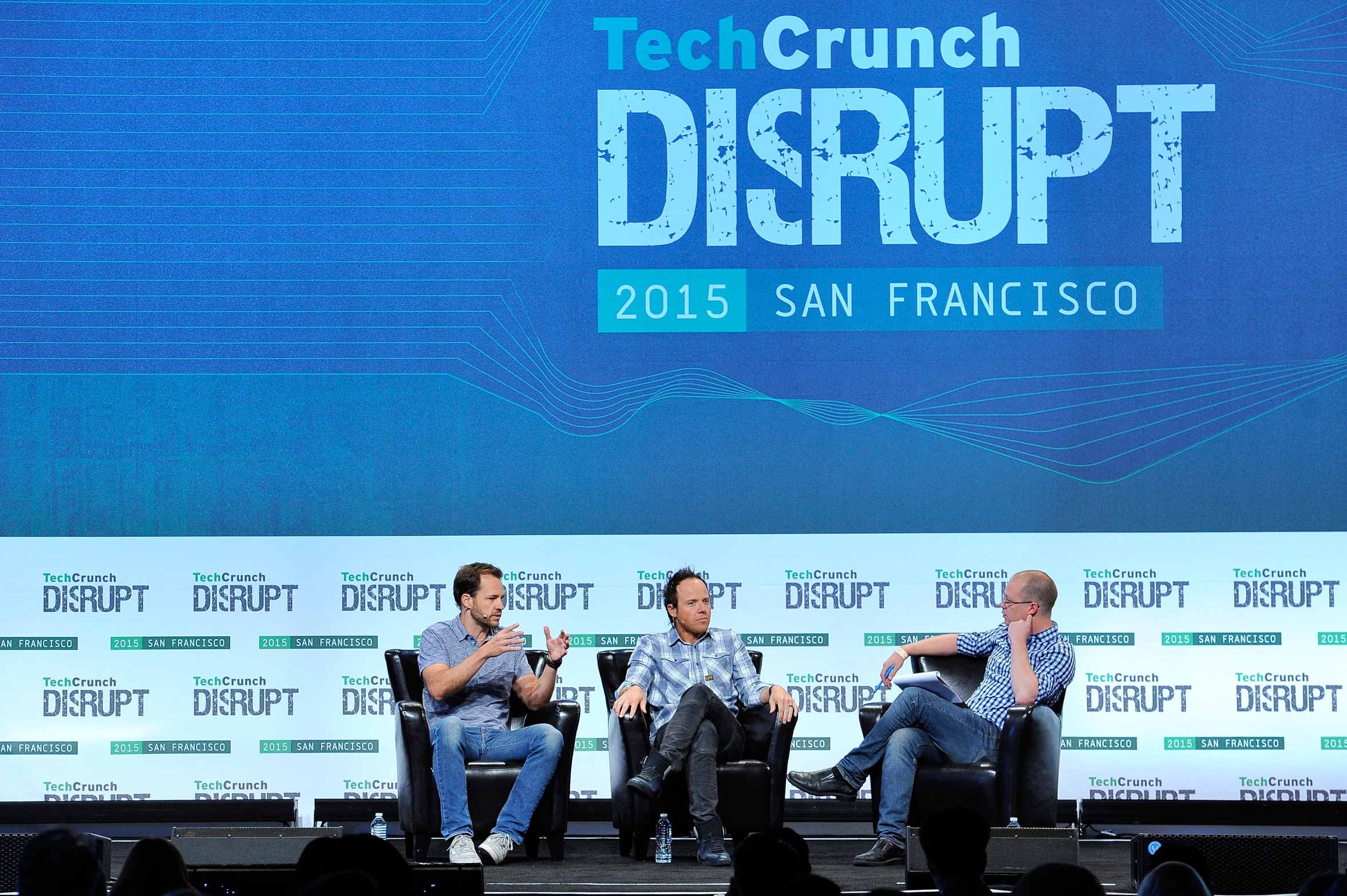 four men sitting on stage in front of a screen and a crowd