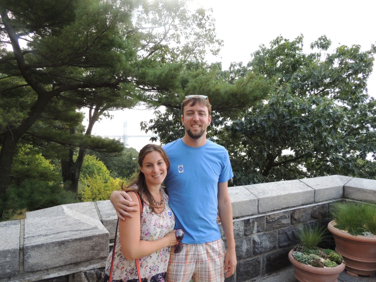 two people standing by a stone wall in front of some trees