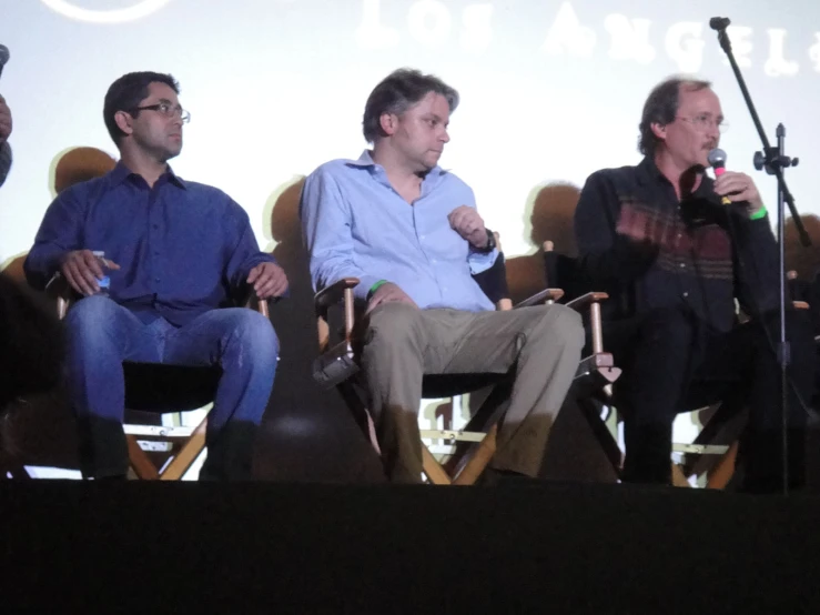 a panel of three men sitting in chairs talking on the stage