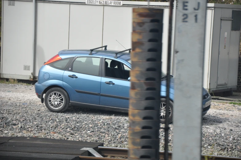 the car is blue on the gravel and has an open trunk