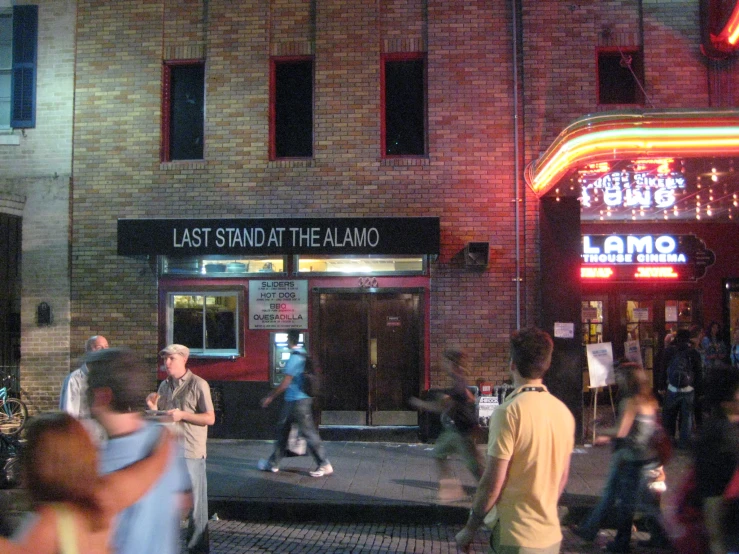 people in the street of an area with lights on and a red sign that reads last station of the alamo