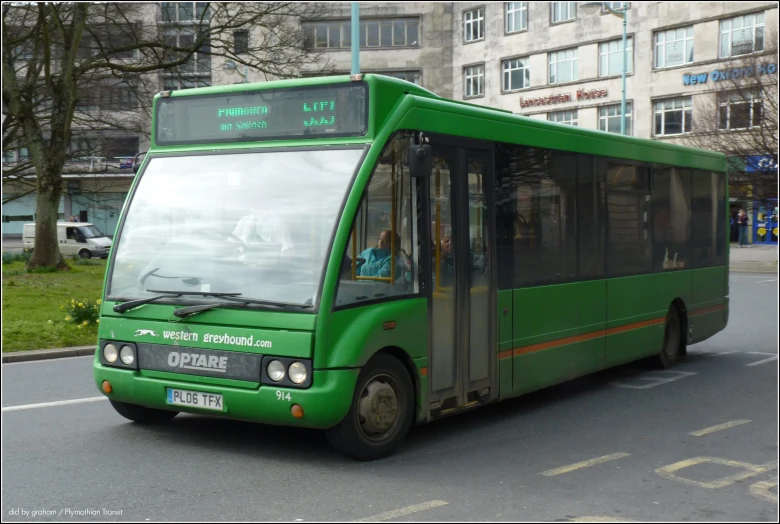 a green bus in front of buildings, and people are sitting inside