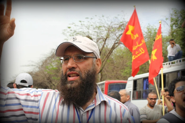 a man with a beard, a baseball cap and glasses stands in front of a group of people