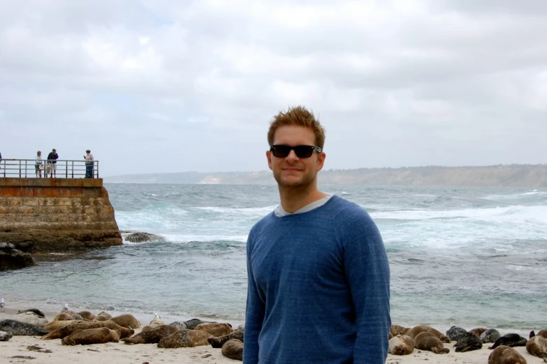 man wearing shades standing on beach by ocean with rocky shoreline