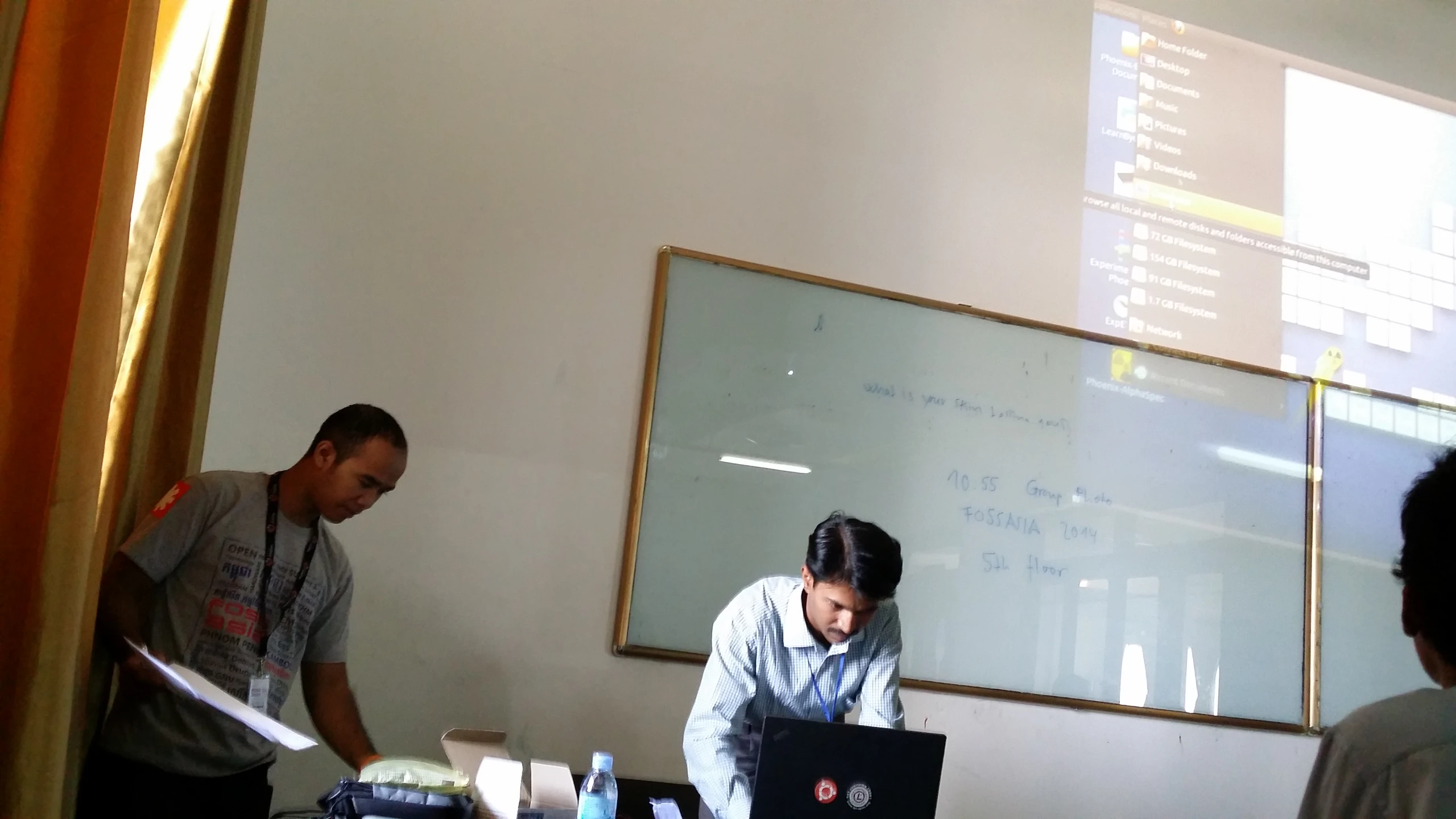 three people with headphones stand around a table, working on their computers