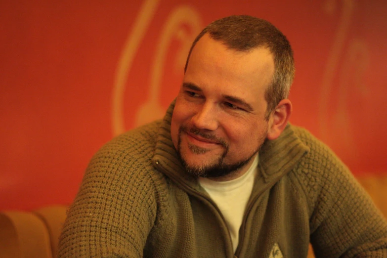 a smiling man sitting at a table with a red sign in the background