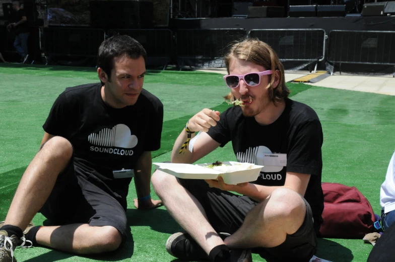 two men with sunglasses on, one sitting down while the other eats some food