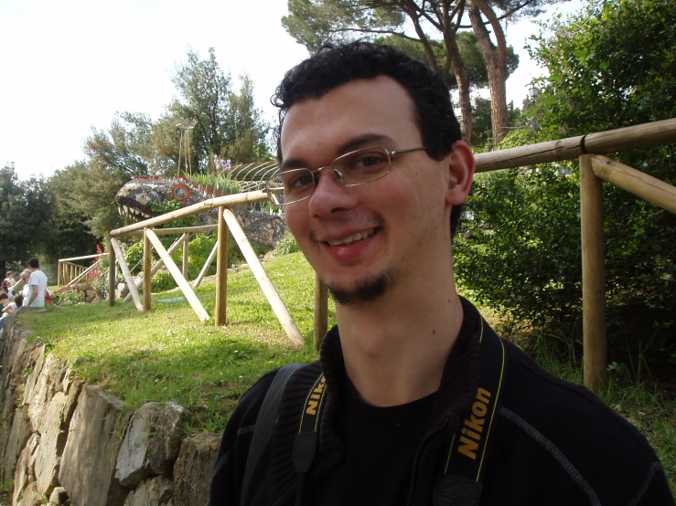 a man with glasses standing near the grass in front of a park