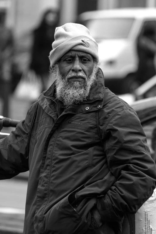 an old man in a jacket and hat leaning against a wall