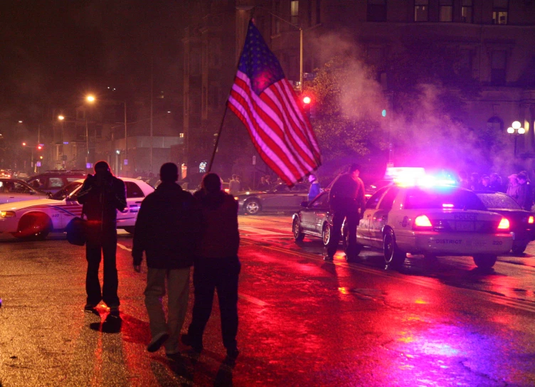 people stand in the street near a car with a american flag