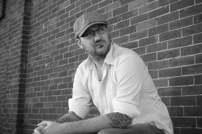 man with glasses leaning against brick wall wearing hat