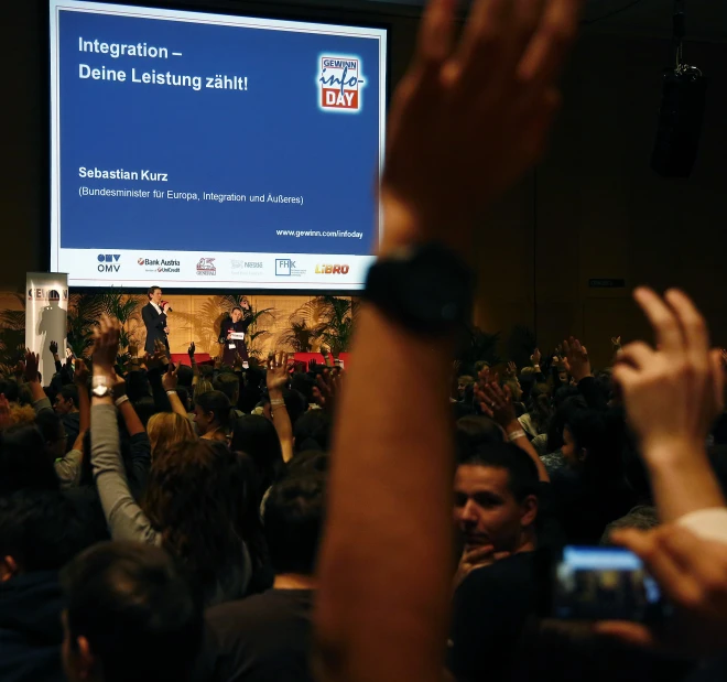 large group of people at a social gathering in a conference hall with an electronic screen displaying information to the audience