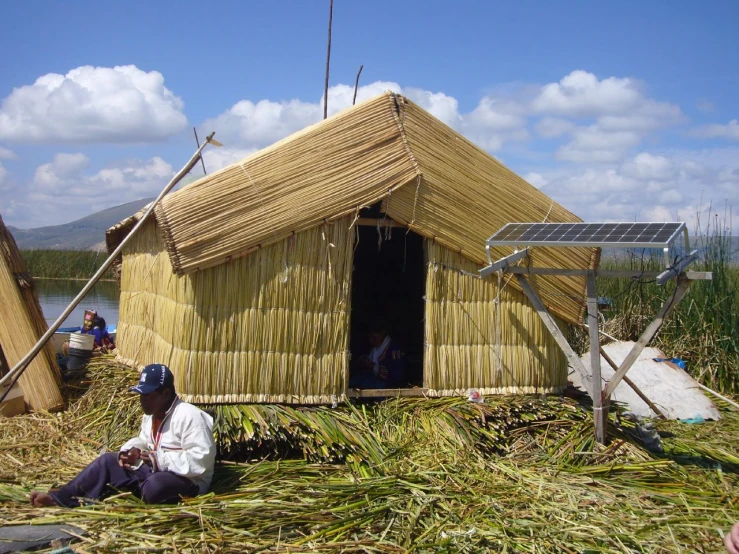 people sitting on straws in front of a hut with water and sky in the background