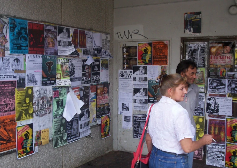 a man and woman walking near a wall covered with newspaper