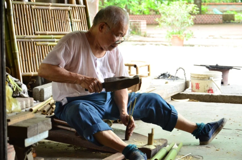 an elderly man sitting on a wooden bench and using a hammer