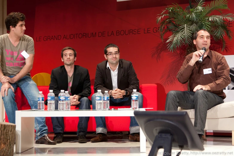 four people sit on red chairs in front of a man with a microphone