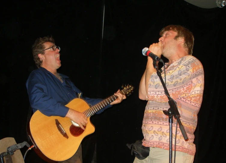 two men singing into microphones with an acoustic guitar