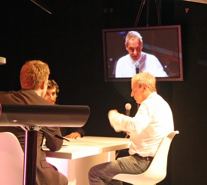 a man and two s sit at a white desk in front of a video screen