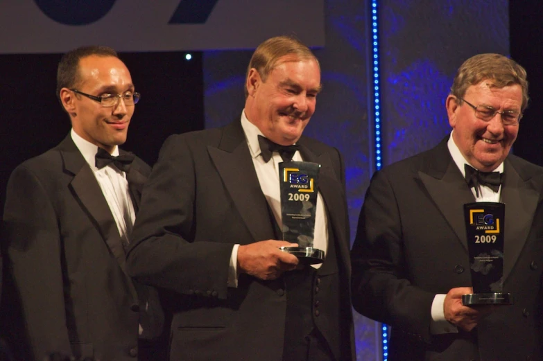 three men wearing black tuxedos standing in front of a screen