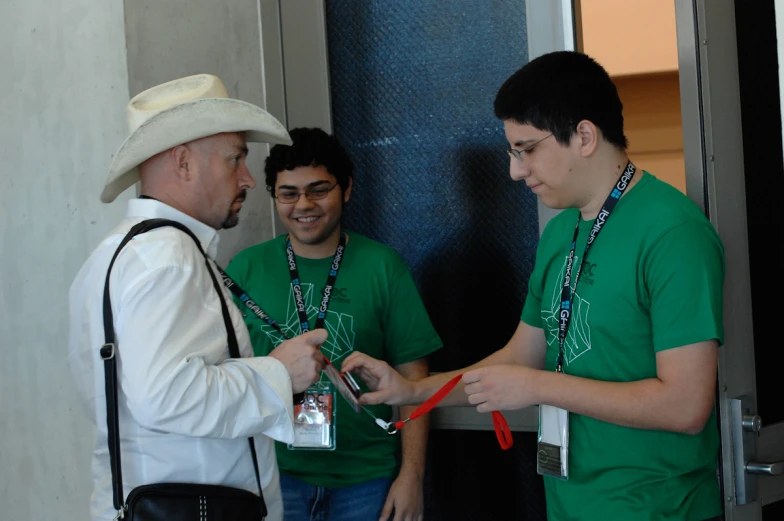 three men with green shirts stand looking at their cell phones