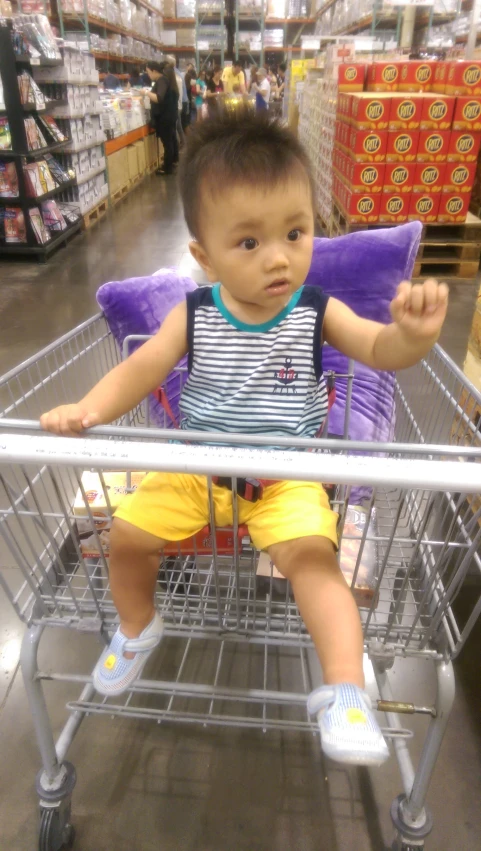 a little boy in yellow shorts sitting in a cart in the store